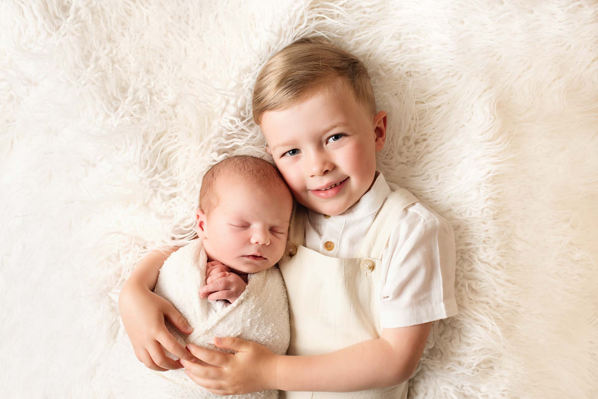 Big brother giving his newborn baby brother a cuddle lying on a fluffy white blanket