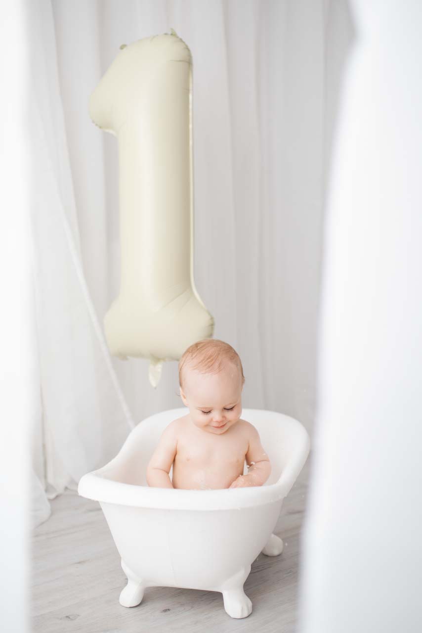 One year old boy sitting in a white roll top bath with a number one balloon behind him