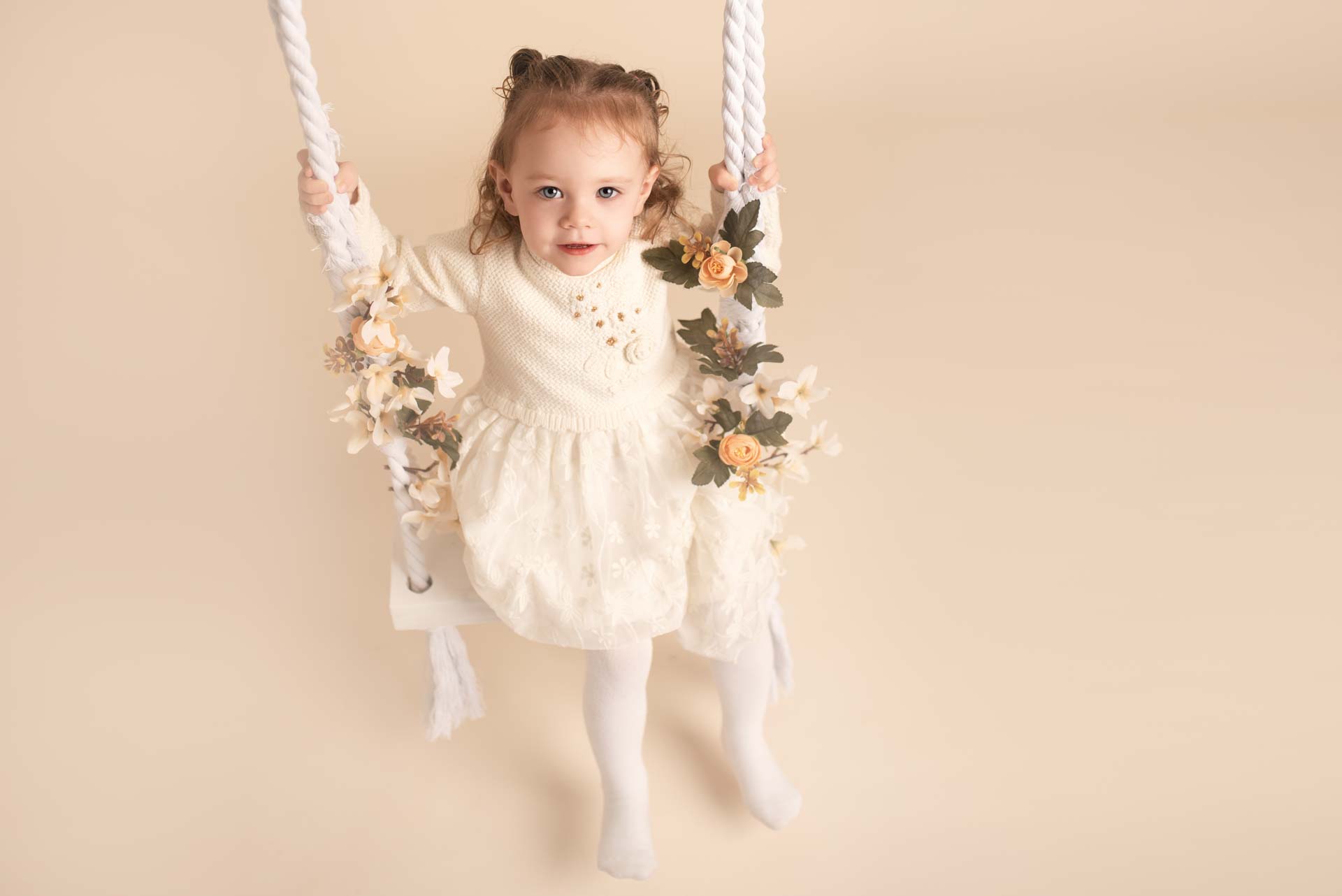 Two year old sits on a wooden swing decorated with flowers at her birthday photoshoot