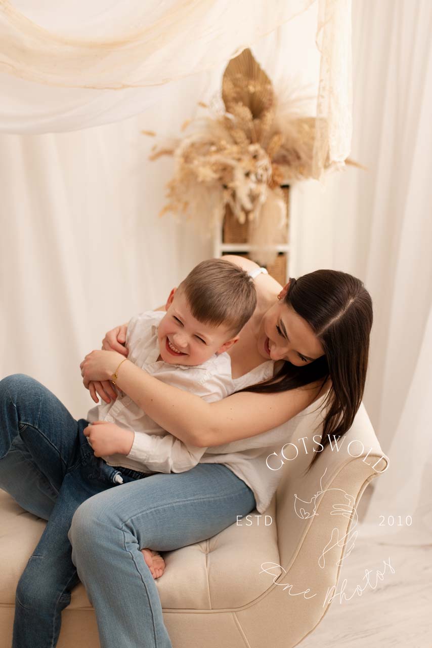 Mummy playing with her son. They are sitting on a chaise and she is tickling him