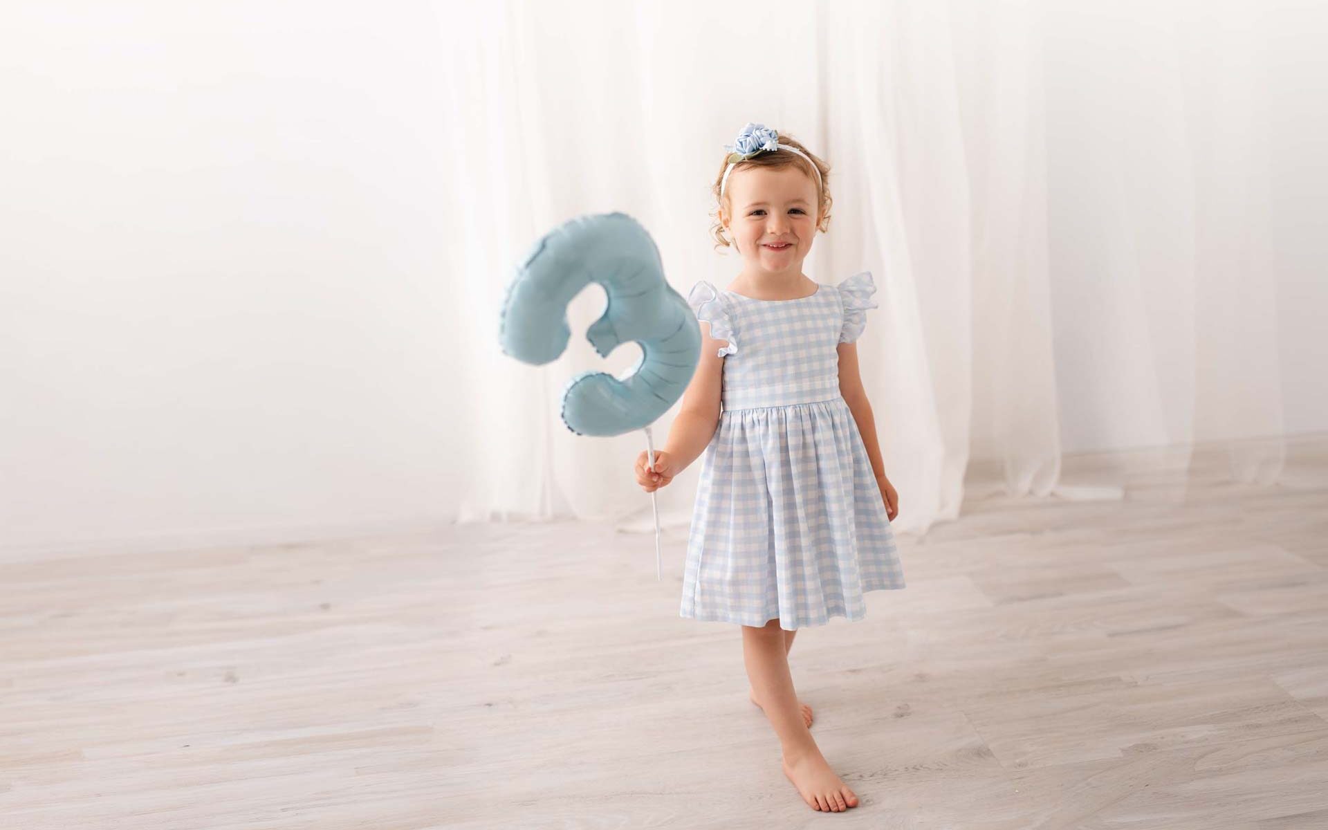 Birthday girl in blue and white checked dress holds a blue number three balloon