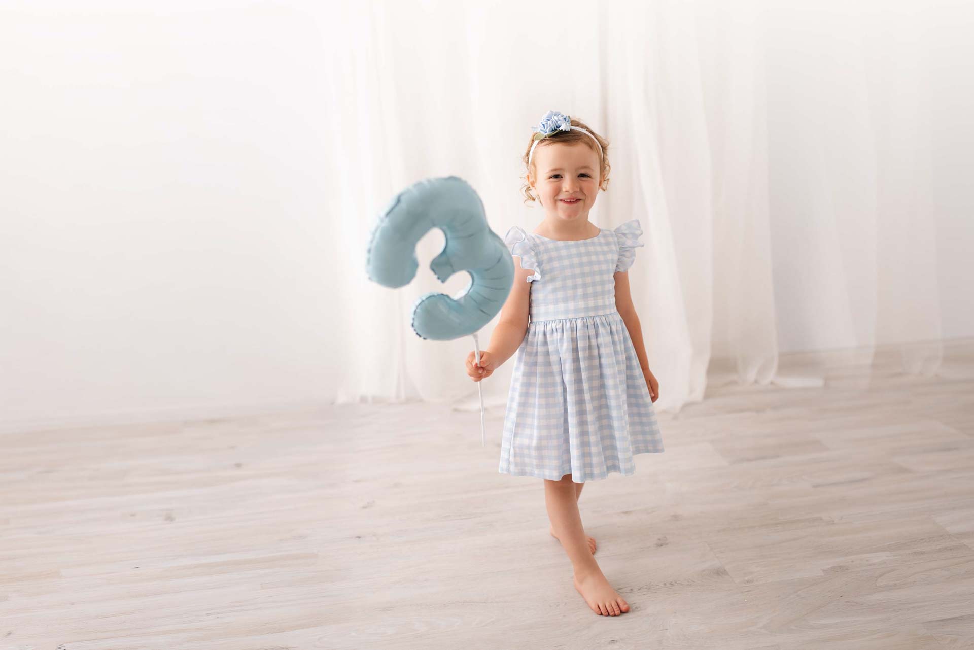 Birthday girl in blue and white checked dress holds a blue number three balloon