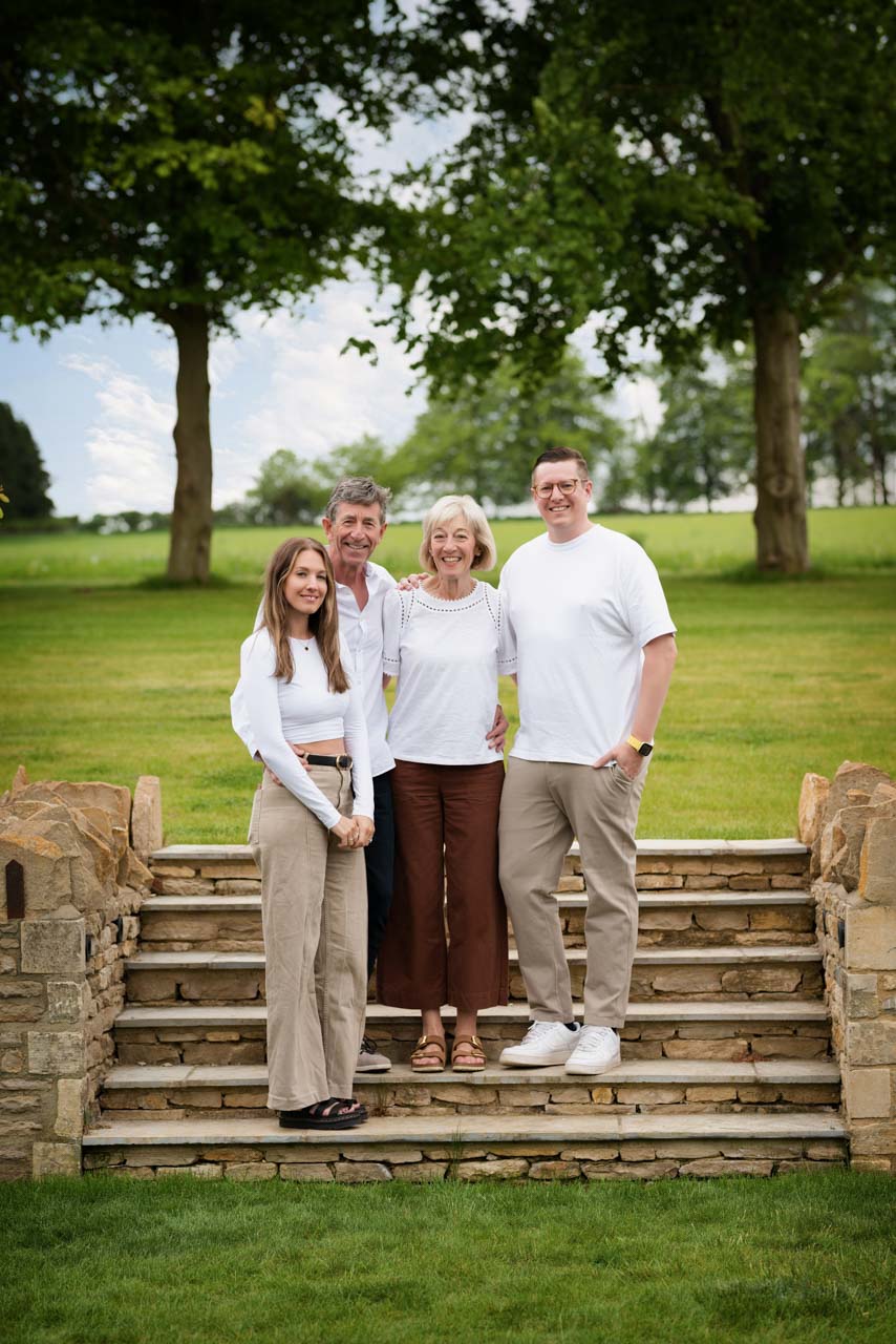 Four members of a family all in white tops and neutral coloured trousers pose on some steps with trees in the background for a 60th birthday celebration