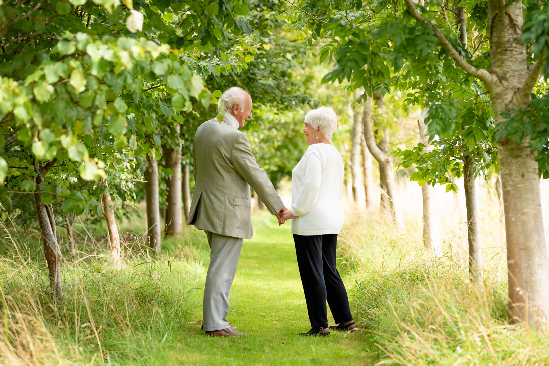 A couple take a walk in the woods. They have stopped to look at each other. The photo is being taken to celebrate a 70th birthday.