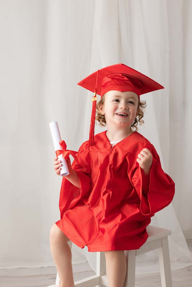 Child celebrates graduating from nursery wearing traditional red gown and hat. Her diploma is in her right hand