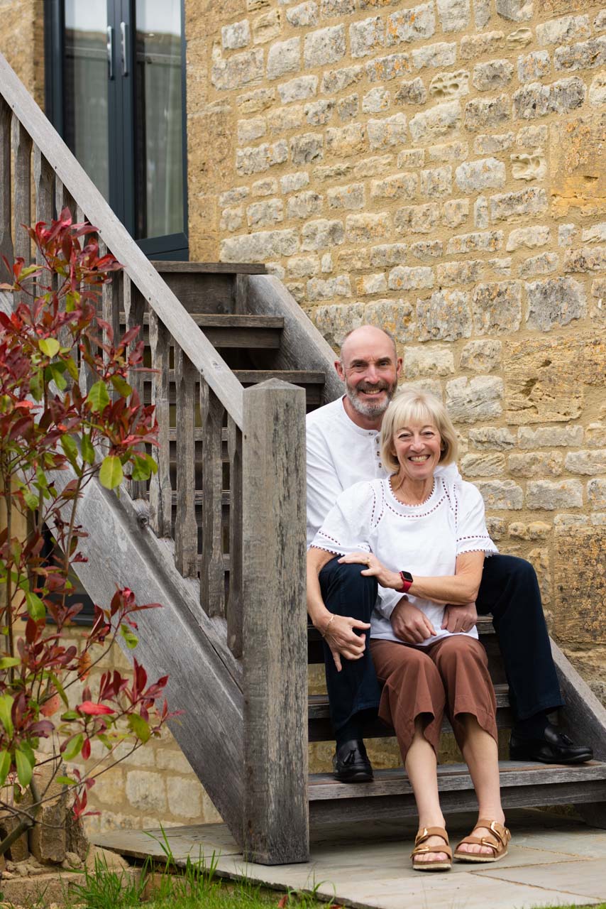 A couple pose on a flight of wooden stairs outside a stone building for photos to celebrate a 60th birthday