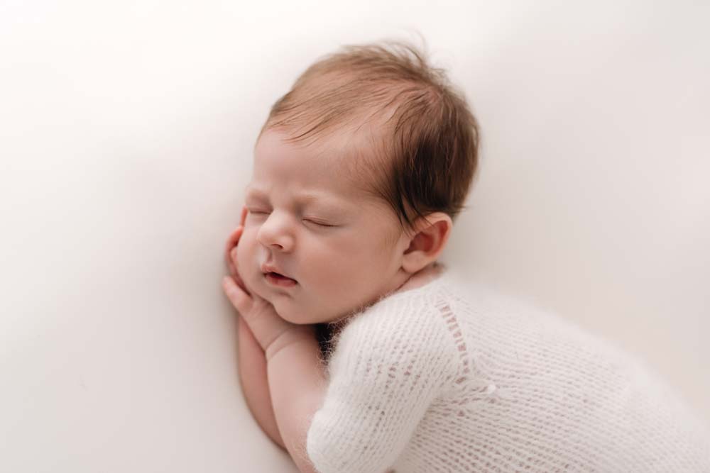 Close up of newborn baby boys head. His hands are under his head and he is wearing a white romper