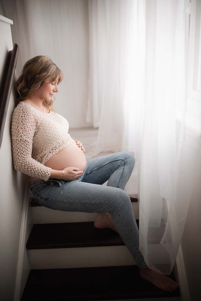 Pregnant women wearing blue jeans and a crochet top sits at the top of a flight of stairs
