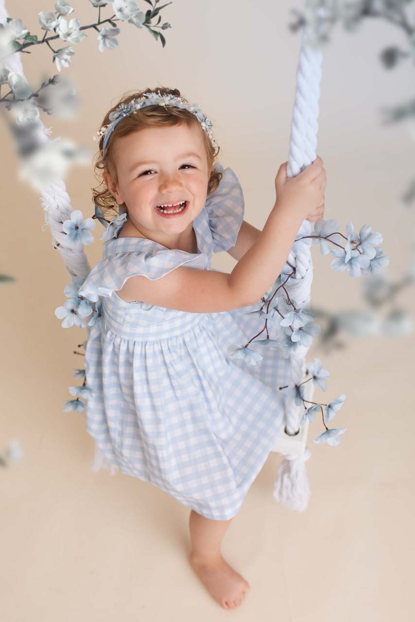 Girl in a blue and white gingham dress sits on a rope swing surrounded by blue and white flowers