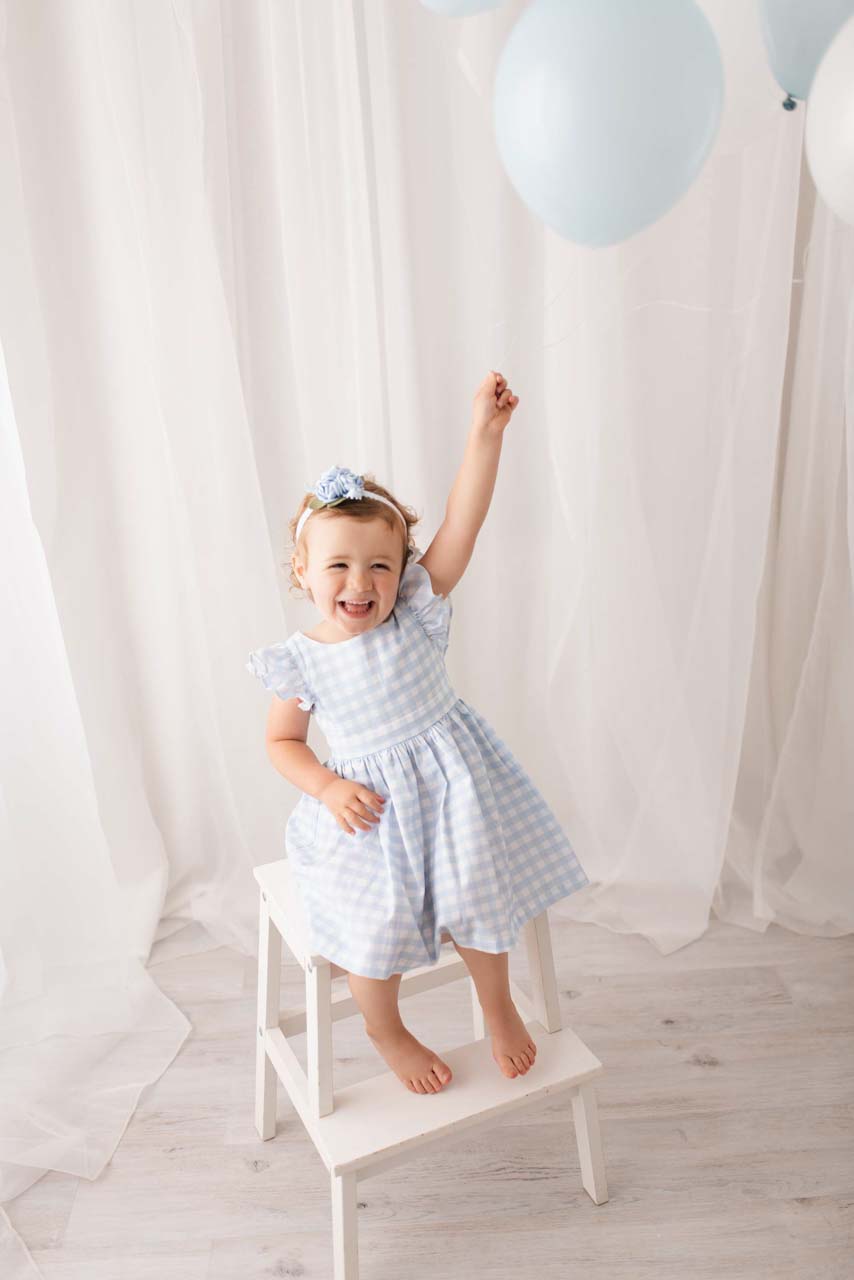 Three year old girl wearing gingham dress sitting on a white stool holding a blue balloon aloft
