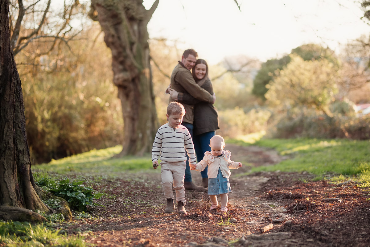 Brother and sister toddle along as mummy and daddy cuddle in the background