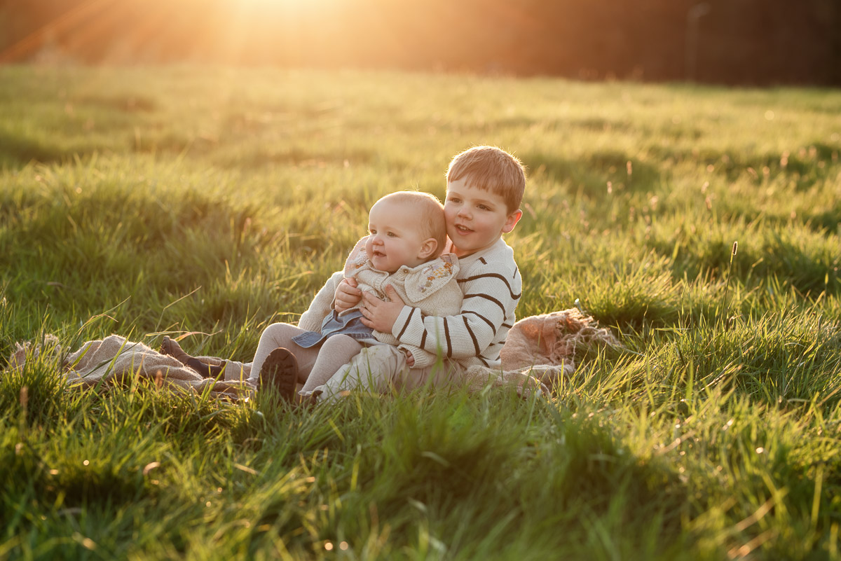 Siblings cuddling in the grass as the sun goes down behind them during their family photography session in Oxfordshire