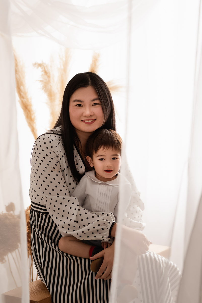 Mother cuddles her son sitting on a wooden bench behind some net curtains