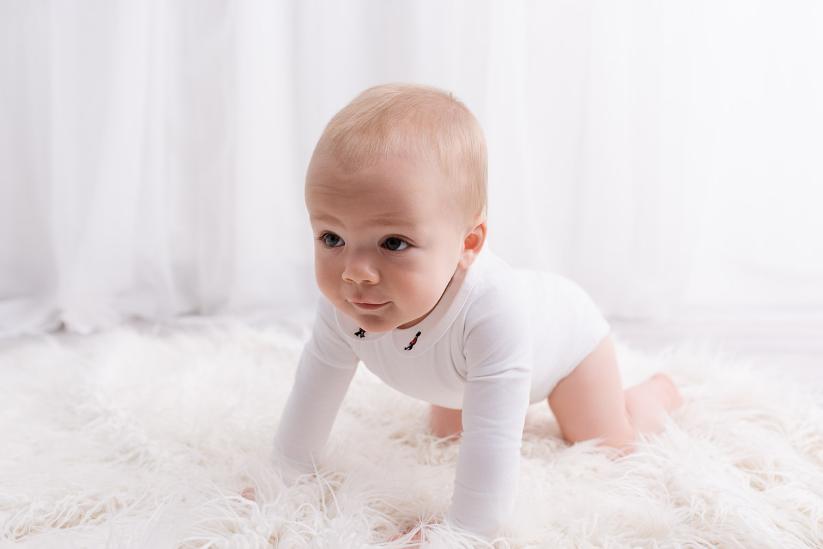 Young boy wearing all white is on all fours on a white fluffy blanket during his photoshoot