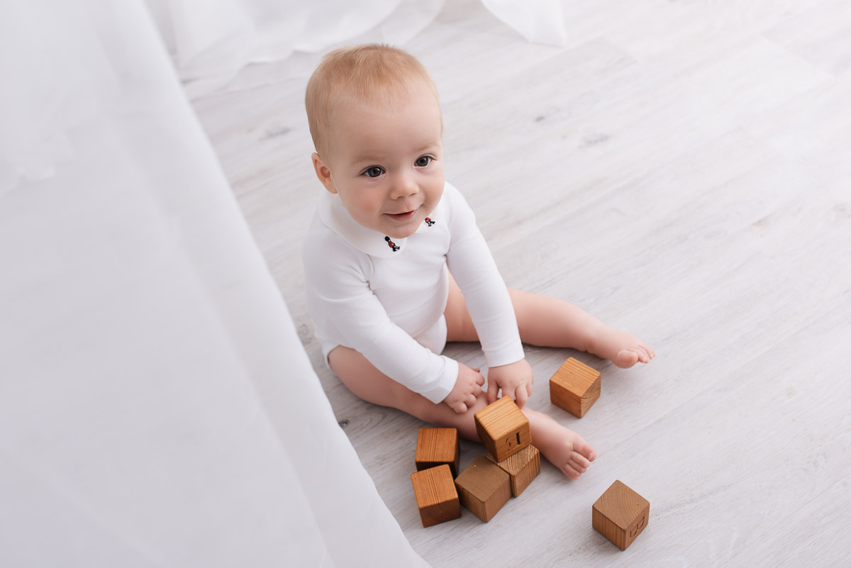 Baby boy wearing white sits playing with wooden blocks