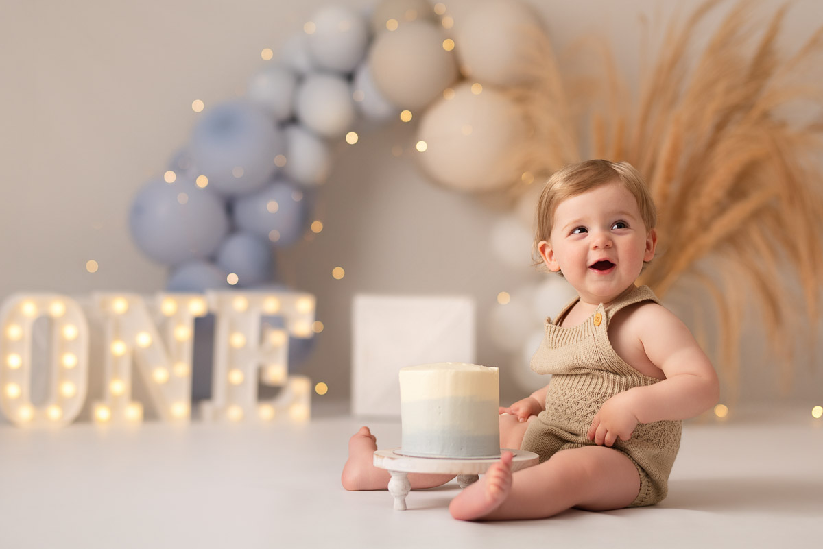 One year old boy gets ready to smash his cake whilst smiling for the photographer in their Oxfordshire studio