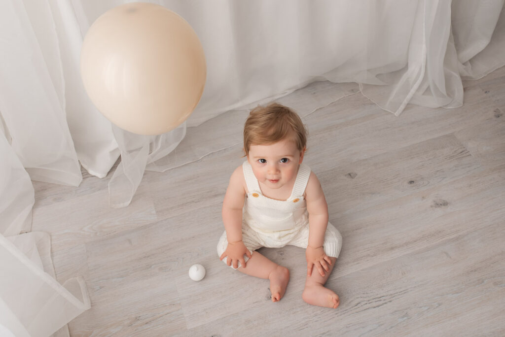 Boy sits on the floor looking up at the photographer. There is one balloon floating in image