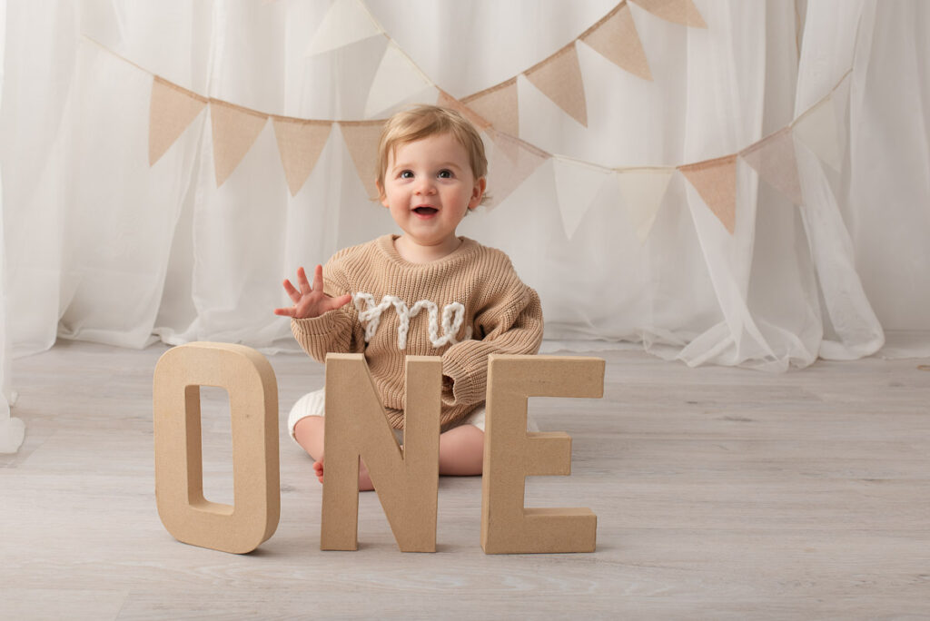 Baby boy is wearing a jumper with the word one on it. He is sitting behind large cardboard letters which also say one