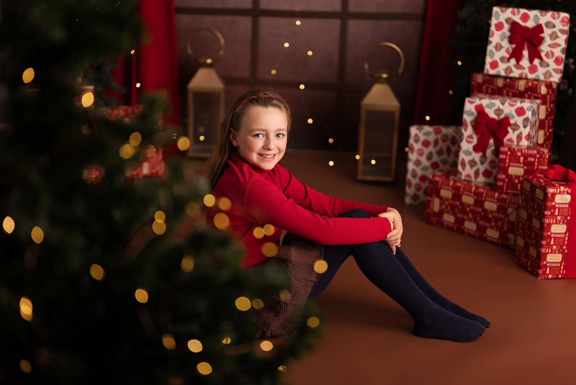Girl in red top sits in front of gold lanterns and wrapped Christmas presents with a Christmas tree in the foreground