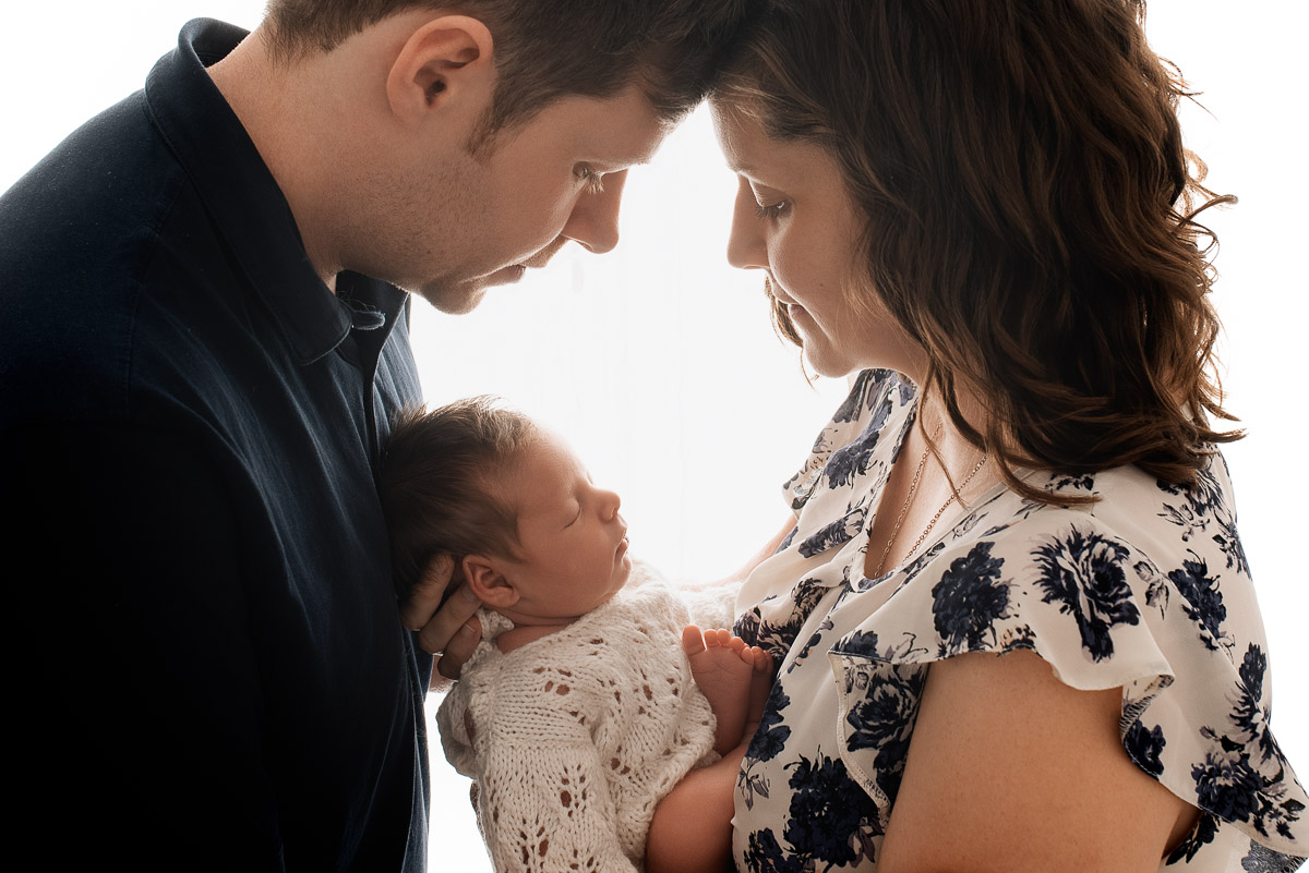 Mum and dad face each other with their newborn daughter nestled between them. The light is coming from behind them