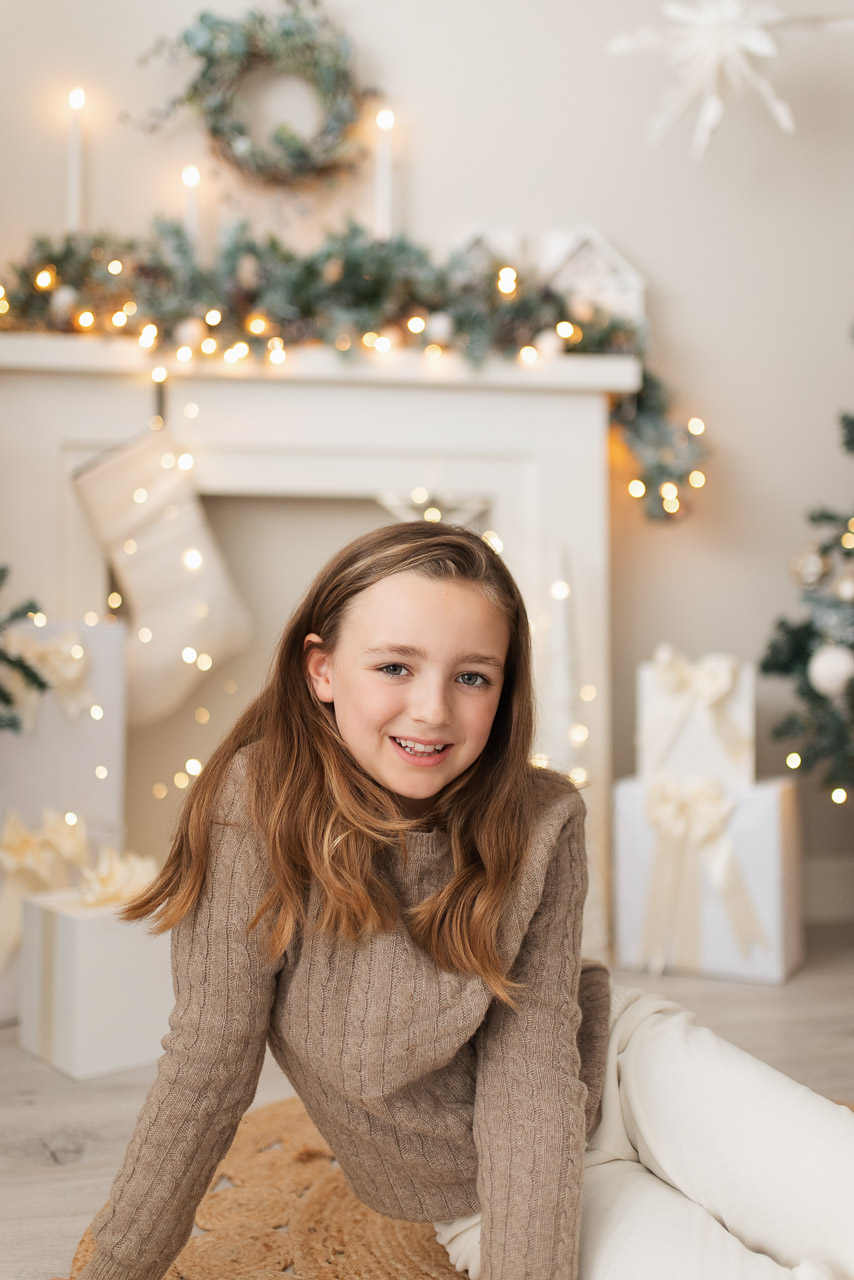 8 year old girl wearing a beige top and white trousers sits in front of a fireplace decorated with candles and Christmas decorations