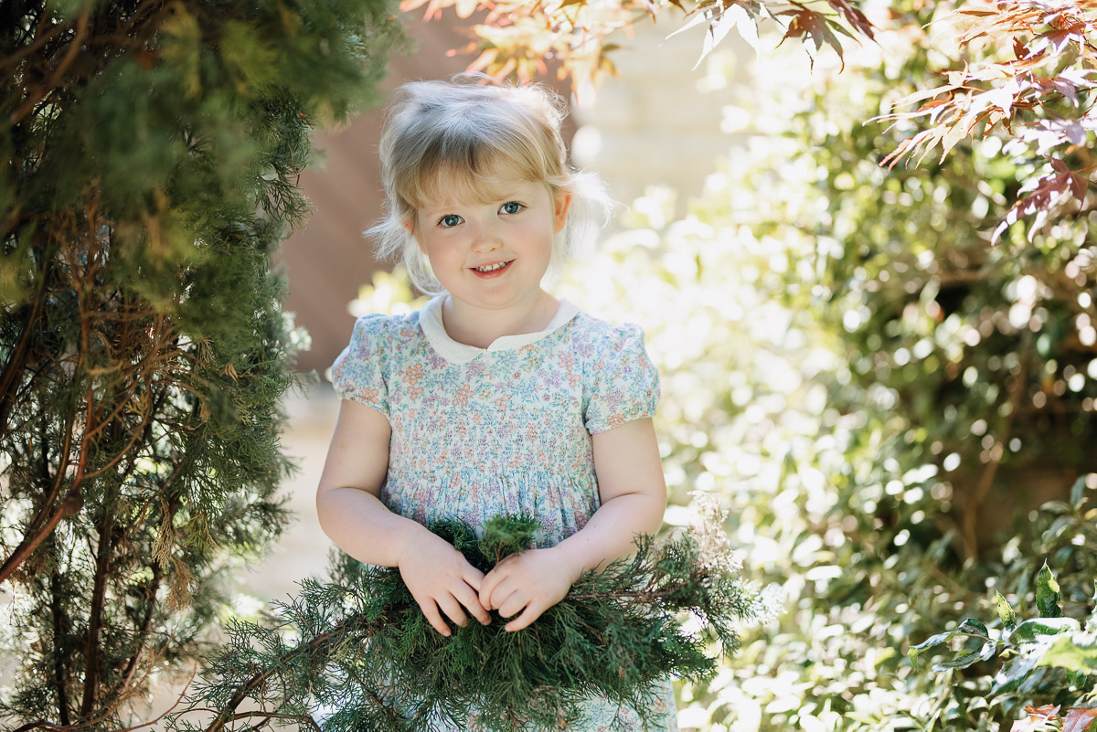 Girl smiles for the camera. She wears a floral dress and is surrounded by evergreens