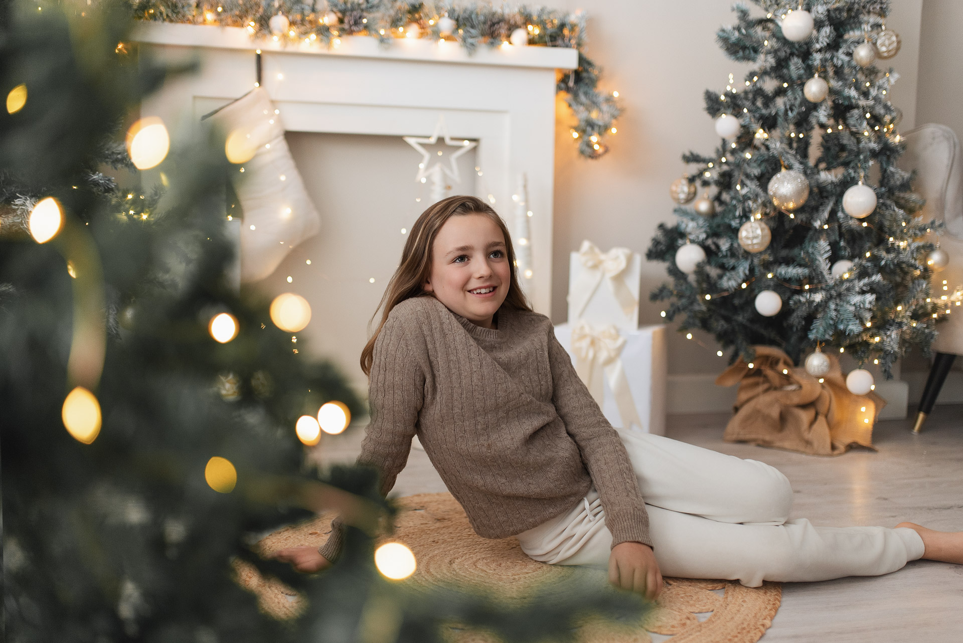 Girl sits in front of fireplace surrounded by Christmas trees for her photoshoot