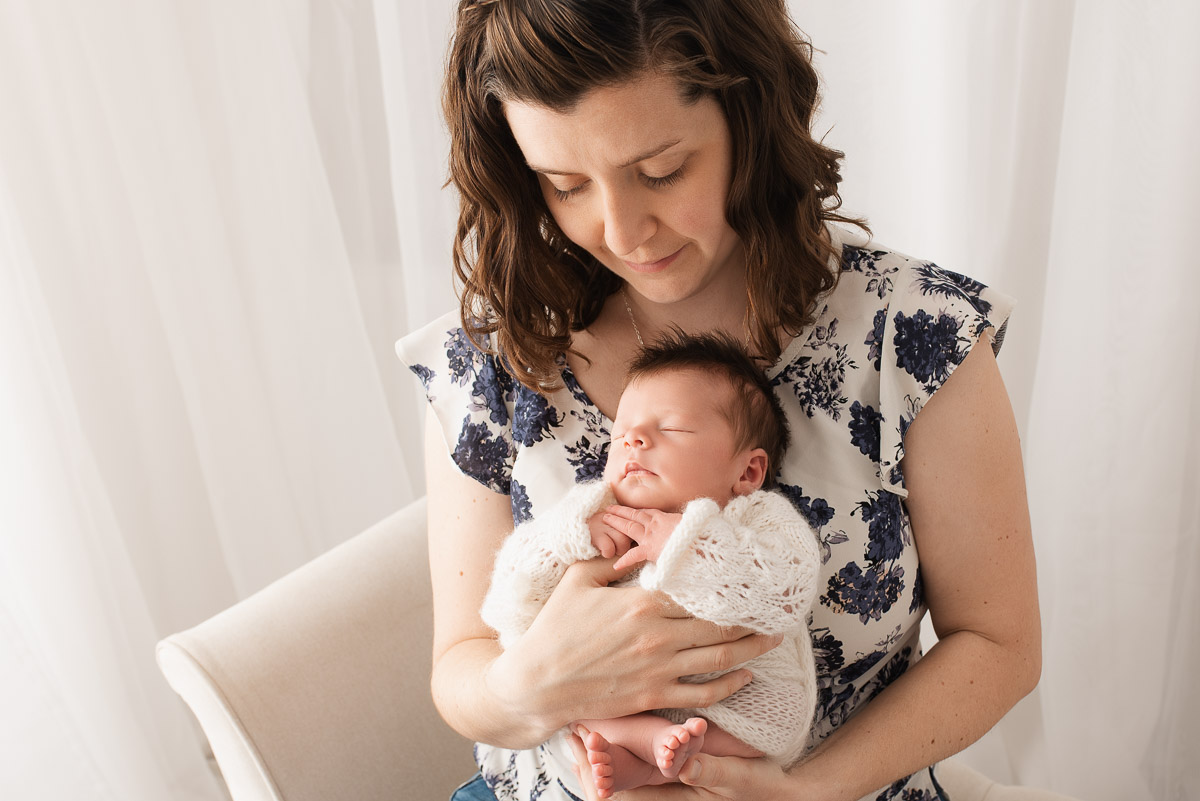 Mother wearing a blue and white dress holds her newborn baby who is wearing white