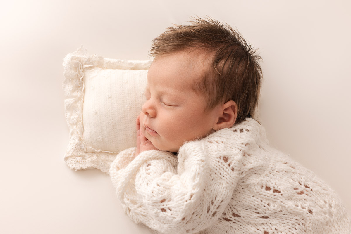Newborn baby girl dressed in white with her hands under her cheeks sleeps on a delicate white pillow