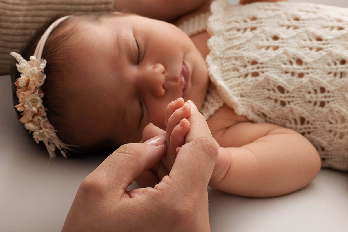 A baby's fingers are curled around her mothers fore finger. The baby is asleep in the background