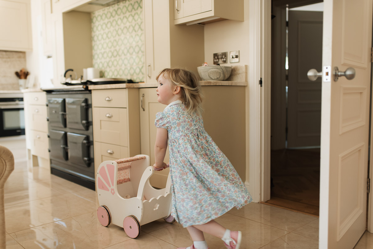 Girl pushing her toy pram around the family kitchen with Aga and worktops in the background
