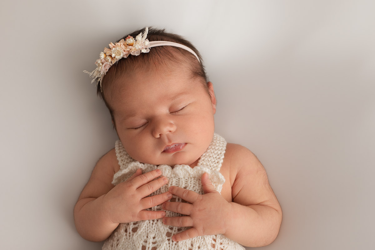 Newborn baby girl wearing knitted romper lies on her back with her hands on her chest. She is wearing a light pink floral headband