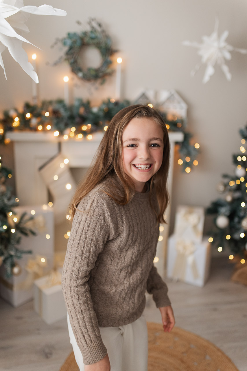 Girl smiles for the camera wearing beige jumper and white trousers with festive fireplace in the background