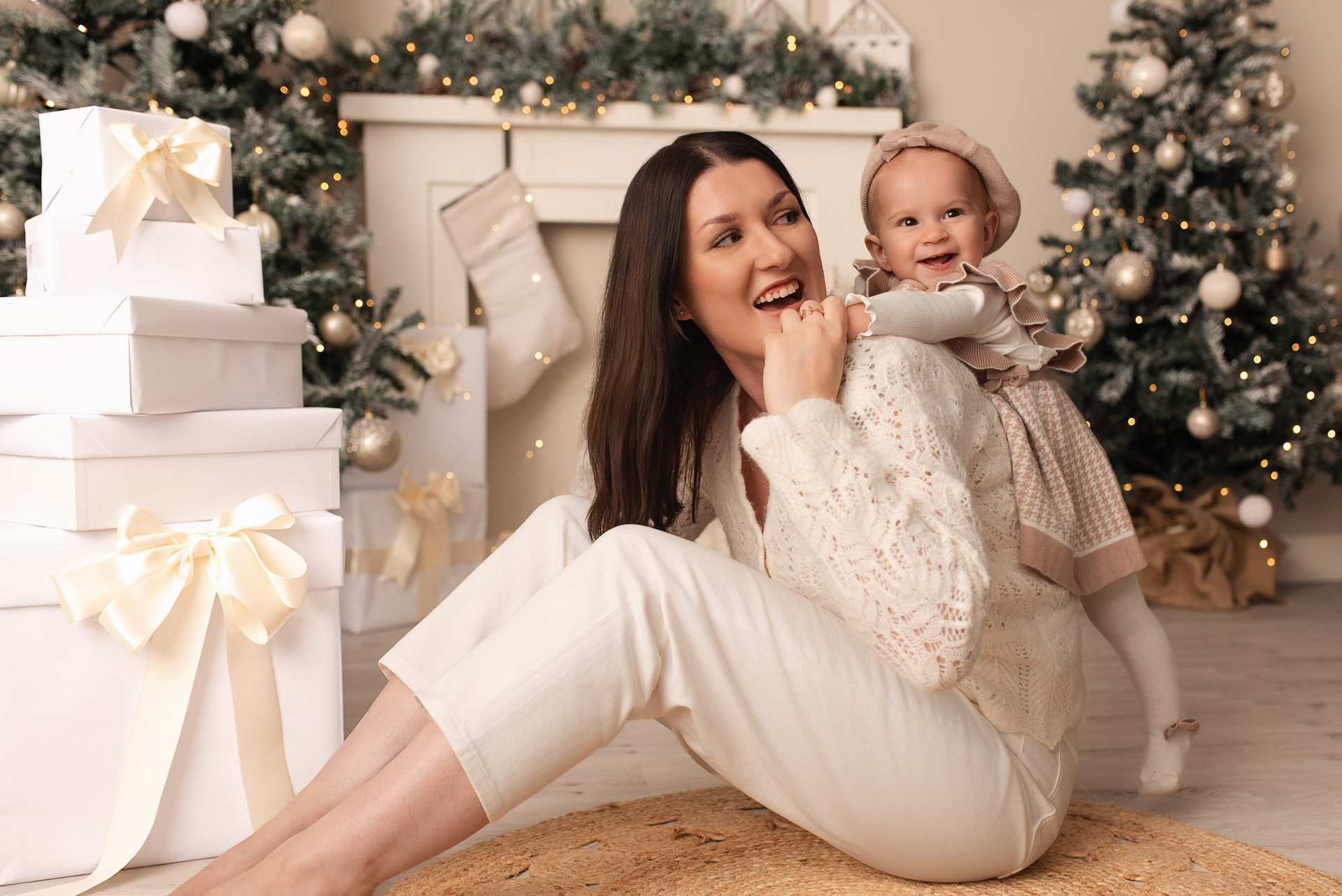 Baby girl climbs up her mothers shoulders. Mummy is sitting on the floor next to a pile of white presents in front of the studio Christmas set up